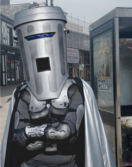 Count Binface. Head and face covered by a bin looking helmet, dressed in grey superhero attire. Arms folded.